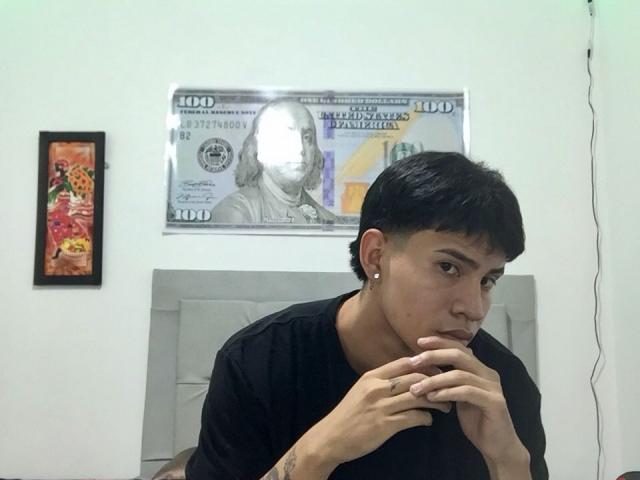 Logannth with short black hair, black shirt, and earrings, looking down with hands near mouth, in a room with a framed dollar bill and colorful picture on the wall.