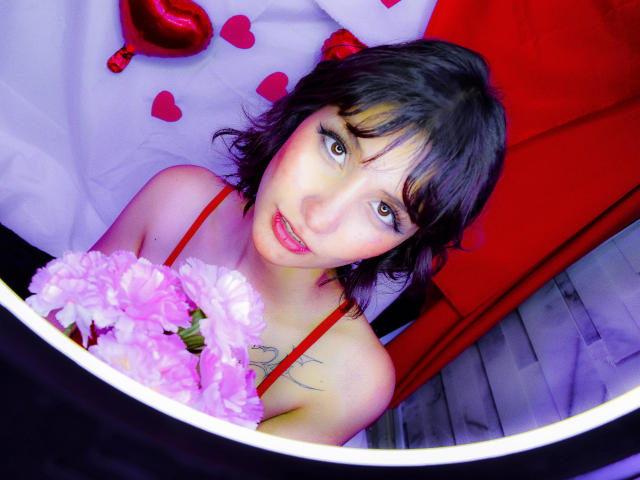 IsabellaHallad with short black hair, red spaghetti straps, pink flowers, tattoos, and a cute face, looking up with a slight smile, in a brightly colored, red and white setting.