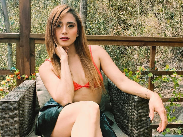 EliBaker with long brown hair, red bikini top, showing small tits, sitting on wicker chair, hand on chin, green eyes, red lipstick, black skirt, forest background.