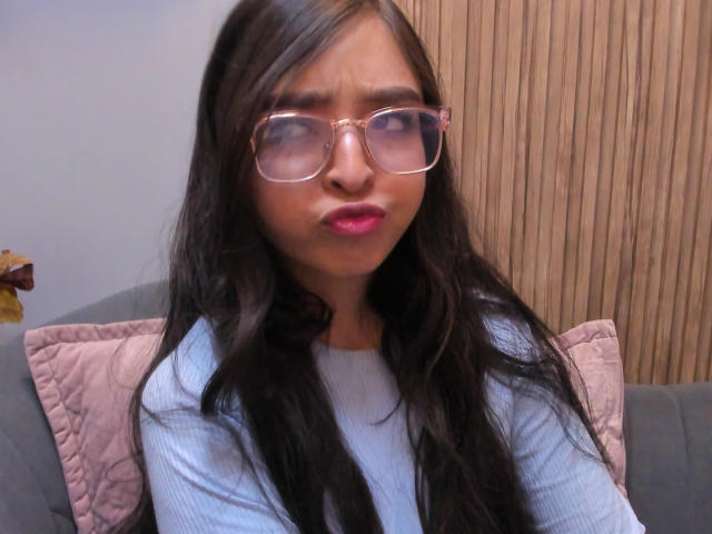 EvaMcAllister with long black hair, pink lips, and clear glasses, wearing a light blue ribbed sweater, making a pouty face, sitting on a gray couch with a pink pillow.