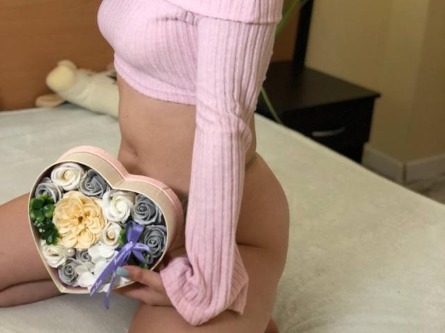 Vailett with pink ribbed long-sleeve top, no pants, holding heart-shaped box with roses and swirled ribbons, sitting on bed, showing bare ass.