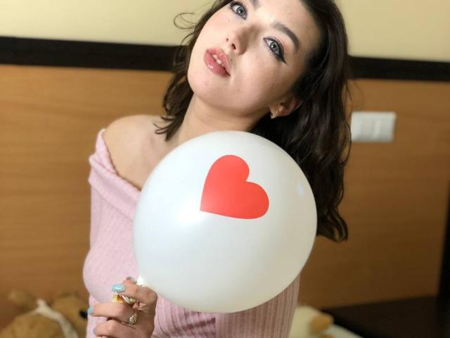 Vailett holding a white balloon with a red heart, wearing a pink off-shoulder top, has dark brown wavy hair, and blue eyes with makeup, in a slightly tilted pose.