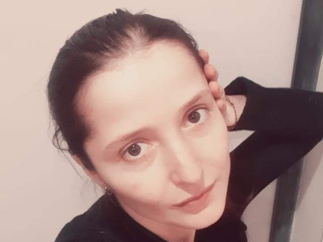 CarenGreen has pale skin, brown eyes, and dark hair in a ponytail. She's wearing a black top, hand on head, and a bracelet, with a slight smile.