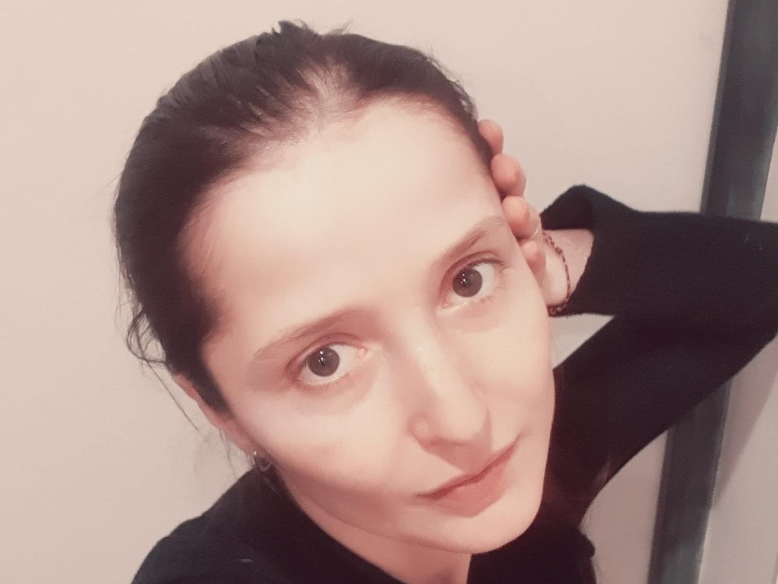 CarenGreen has pale skin, brown eyes, and dark hair in a ponytail. She's wearing a black top, hand on head, and a bracelet, with a slight smile.