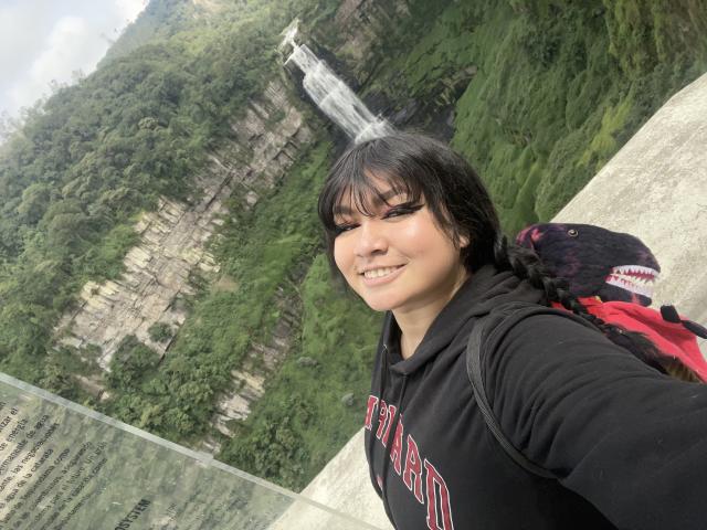 LillyMayer with black hair, wearing a black hoodie with red letters, smiling, holding a red and black backpack, standing near a waterfall in a forest.