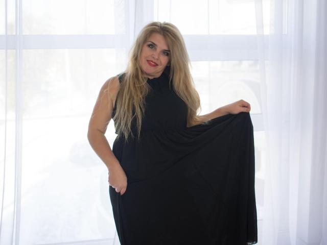 JulyaBailly, con cabello rubio largo, vestido negro sin mangas, sosteniendo la falda, sonriendo, de pie frente a una cortina blanca, con luz proveniente de atrás. Se ven los senos debajo del vestido..