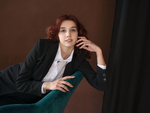 LettyCurly with red curly hair, wearing a black suit and white shirt, leans on a green velvet chair, hand on face, looking slightly bored. Boobs covered by suit.