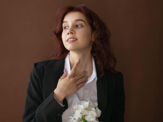 LettyCurly met rood haar, gekleed in een zwart blazer over een wit shirt, met witte bloemen, hand in de buurt van de nek, lachend, bruine achtergrond..