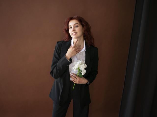 LettyCurly with red curly hair, black suit, white shirt, holding white flowers, hand on neck, looking slightly up, smiling softly, standing against brown wall.