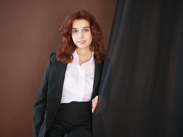 LettyCurly with wavy red hair, wearing a black blazer and white shirt, black pants, smiling, holding a dark curtain on the right, looking relaxed.