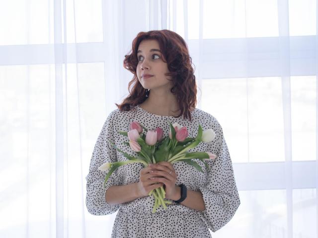 LettyCurly with curly red hair, wearing a white polka dot dress, holding a bouquet of pink and white tulips, looking to the side, wearing a black wristwatch.