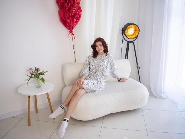 LettyCurly with red curly hair, wearing a white polka-dot dress and white socks, sitting on a white couch, legs crossed, red heart balloon behind her, yellow light on tripod.