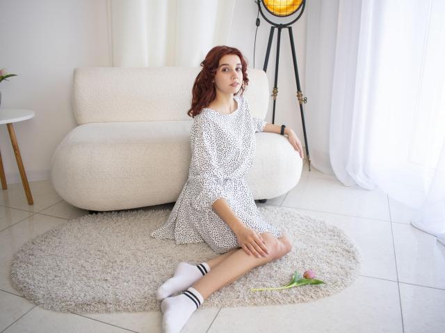 LettyCurly with red hair, sitting on a white rug, wearing a white polka dot dress, white socks, and a watch, with a pink rose on the floor.