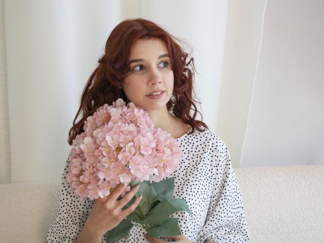 LettyCurly with wavy red hair, holding a big pink hydrangea, wearing a white polka dot shirt, sitting on a white couch, looking to the side with a gentle smile.