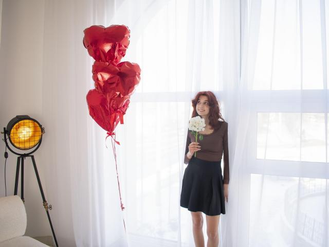 LettyCurly with red balloon cluster, brown top, black skirt, holding white flower, standing by window, next to yellow spotlight. Her ass is slightly visible. Her tits are small.