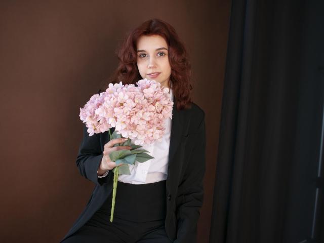 LettyCurly with red curly hair, holding pink peonies, wearing a black suit jacket, white shirt, and black pants, has a cute face, and is looking at the camera.