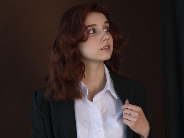 LettyCurly with wavy red hair, green blazer, white shirt, looking to the side, holding her jacket, ring on her finger, soft expression, medium skin, subtle makeup.