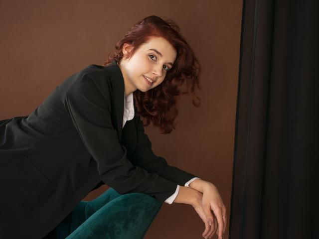 LettyCurly with red curly hair, black suit, white shirt, and teal pants, leaning forward with a smile, hands on knee, looking at camera. Her tits are not visible.