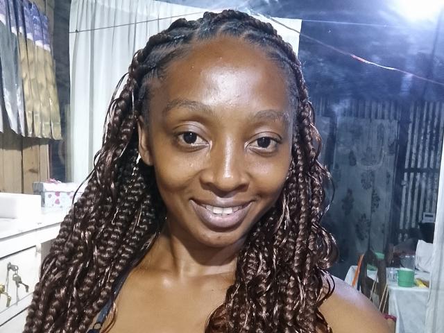 Anjelgas with dark brown, wet, braided hair, smiling, showing small white teeth, shiny brown skin, wearing a black top, in a brightly lit room with a white curtain.