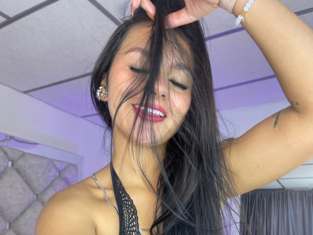 AlessiaFerry, com longos cabelos pretos, sorrindo, olhos fechados, segurando o cabelo, vestindo um top preto de alças, batom vermelho, colar de prata e uma pulseira rosa, mostrando uma tatuagem no braço direito..