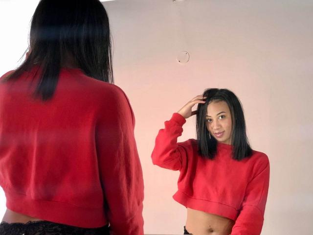 AbbyyRosse with black hair, red crop sweater, black lace underwear, looking at mirror, hand in hair, showing midriff and navel, smiling, reflection of back and front visible.