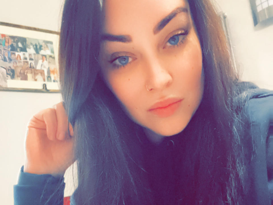 MelinaBella with blue eyes, dark brown hair, and a nose ring, holding her hair, wearing a dark top, looking at the camera with a slight smile.