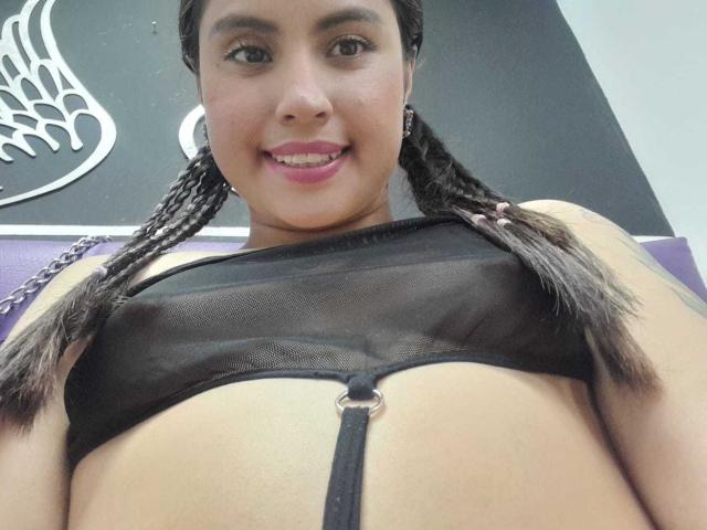 PaulaCeleste with braided hair, wearing a black mesh bra that exposes her tits and a strap that runs down her belly, smiling with pink lipstick.