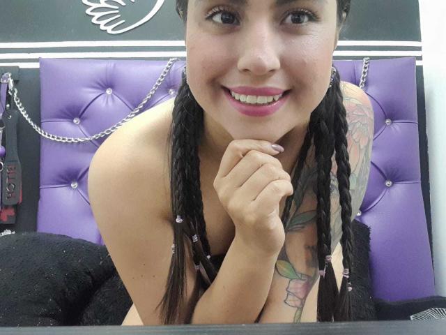 PaulaCeleste with pigtails, smiling, has pink lipstick, tattoo on right arm, and is topless, showing small tits, leaning on a purple tufted headboard with black chains.