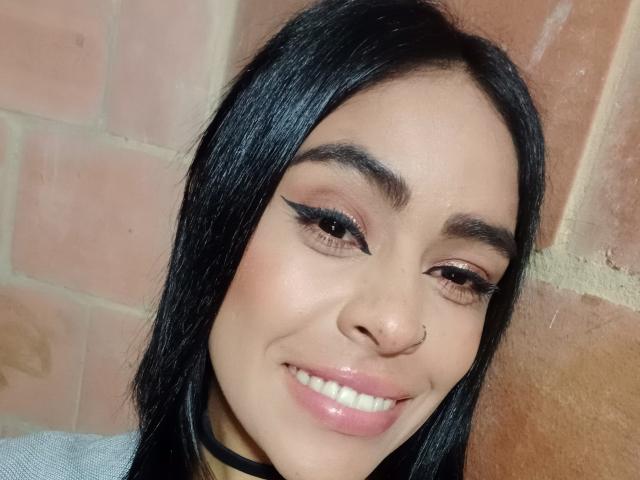 StellaBeckker with black hair, nose ring, and pink lipstick, smiling with winged eyeliner, glossy pink lips, and a cute face, standing against a brick wall.