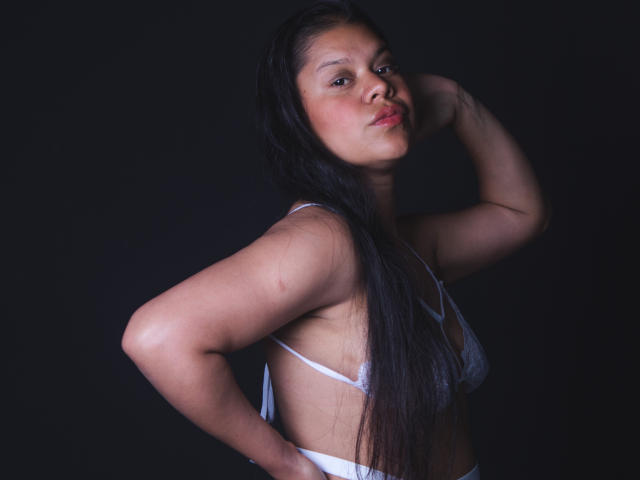 ClaireRouse with long black hair, white bra, and hand behind head, showing her tits and ass, against a dark background. Her face has a slight, confident smirk.
