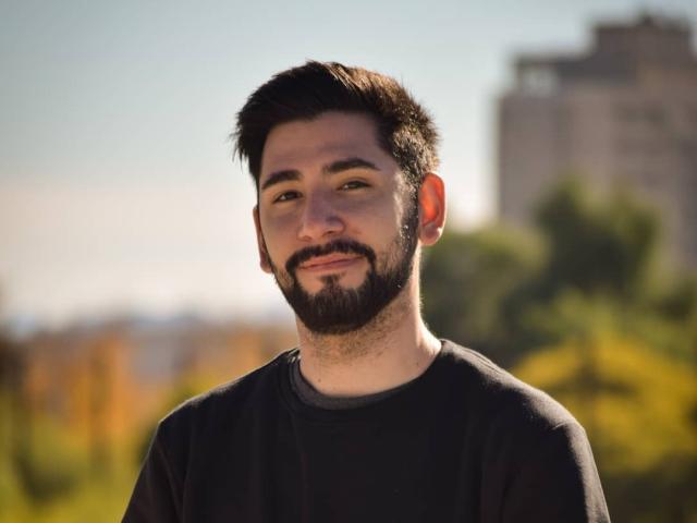 ElMagico has a dark beard, short black hair, and a slight mustache, wearing a black shirt, with a calm, slightly smiling expression in a sunlit outdoor setting.