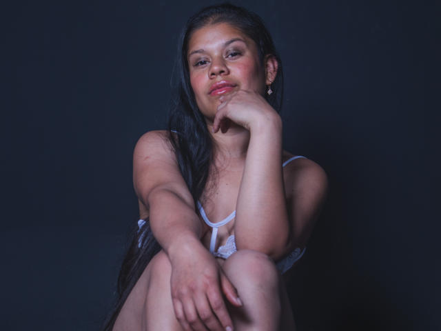 ClaireRouse with long black hair, sitting, wearing white lingerie, resting chin on hand, smiling, hands on knees, dark background, visible tits, soft light, casual, confident, sexy.
