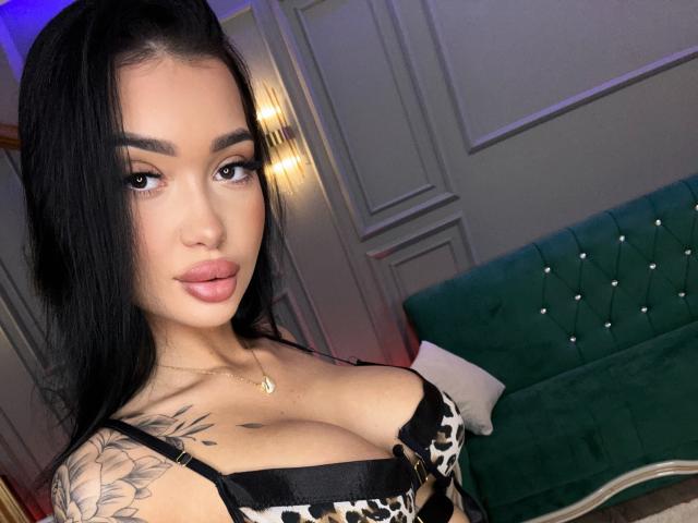 SashaFerrera with long black hair, full lips, and big tits in a black and white patterned top, wearing a gold necklace, and showing a tattoo on her right shoulder.