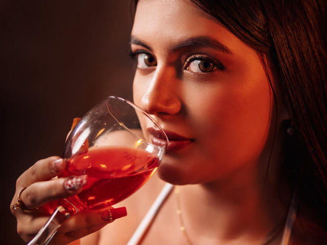 MiaLeiya holding a glass of red wine, slightly tilting it towards her lips, with a subtle, sexy smile. She has brown eyes and long, dark brown hair.