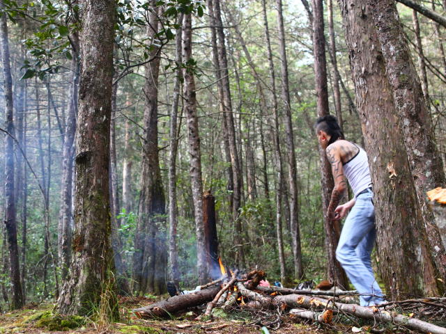 YeremyWalker, com tatuagens, vestindo uma camiseta branca, calças jeans azuis, em pé perto de uma pequena fogueira em uma floresta, cortando lenha, fumaça subindo, toras de madeira no chão, árvores altas ao redor..