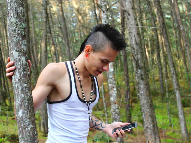 YeremyWalker with a mohawk, white tank top, bead necklace, tattoo on arm, holding phone, leaning on tree in forest. Looking down, slightly smiling. Muscular, fair skin, red ear.