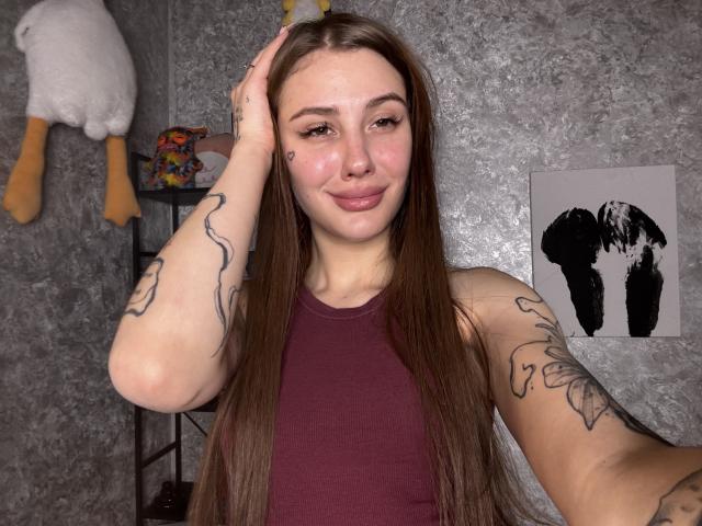 SensualMila with long brown hair, smiling, wearing a maroon tank top, showing tattoos on arms, hand in hair, soft pink lips, fair skin, in a gray textured room.