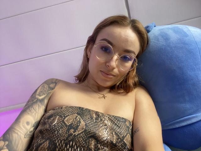 SarahRousen with glasses, brown hair, snake-pattern dress, tattooed arm, laying back on blue pillow, slight smile, purple light, gold necklace, upper body shot, relaxed.