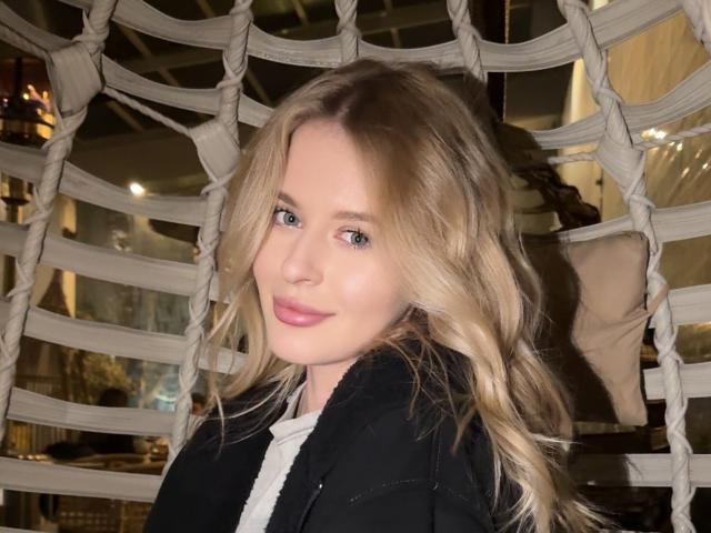MarryMii with blonde hair, blue eyes, pink lips, black coat, and white shirt, standing in front of a rope-patterned wall. Her tits are small, she's cute.