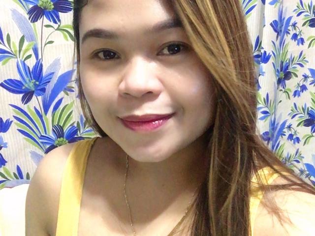 WildParadisePh, with brown hair, smiling, wearing a yellow top, has glossy pink lips, and a thin necklace, showing her cute face and soft skin.
