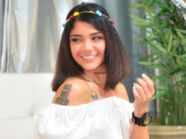 TrinaHarpper, with medium brown skin, black shoulder-length hair, and a colorful headband, smiles, showing off her white off-shoulder top, tattoos, and gold necklace, holding her manicured hand.