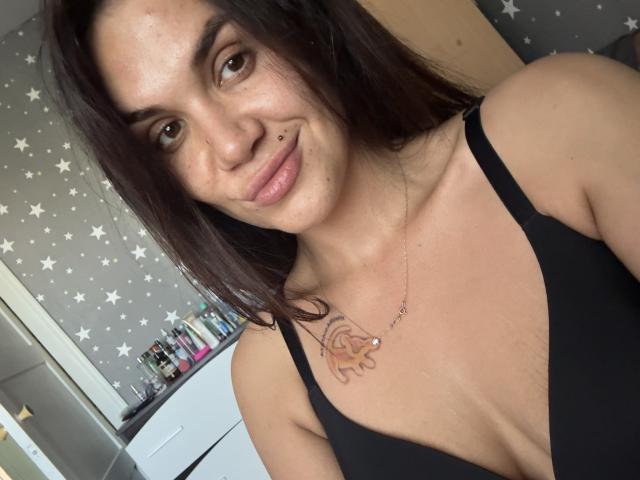 SashaWild, con cabello oscuro, un piercing en la nariz y una camiseta negra, mostrando sus pequeños senos y un tatuaje de un sol en el pecho, sonriendo a la cámara..
