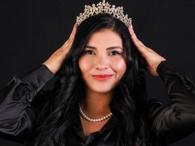 SkyblueKate with long black hair, wearing a silver tiara, black satin shirt, and pearl necklace, hands on head, smiling, red lipstick, dark background.