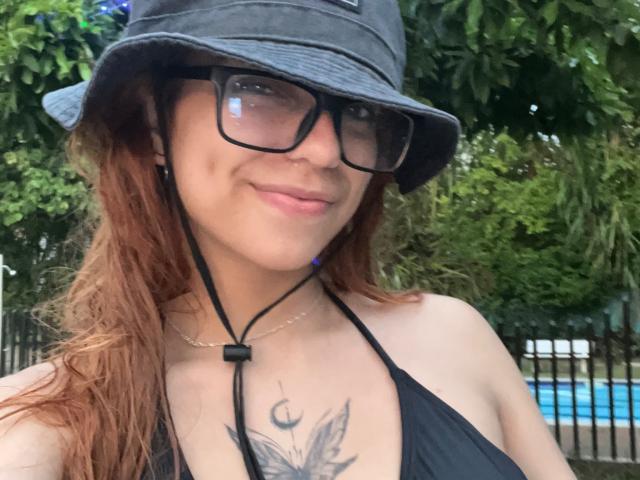 SamantaCarter with red hair, black glasses, and bucket hat, wearing a black halter top, showing a tattoo of wings on her chest, smiling, with earbuds.