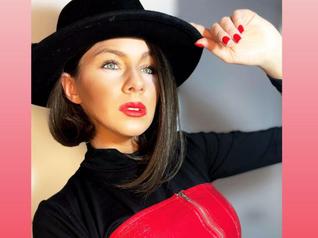 MiSSDiane with red lipstick, black hat, and red zipper dress, holding hat with red nail polish, showing off her tits and ass.