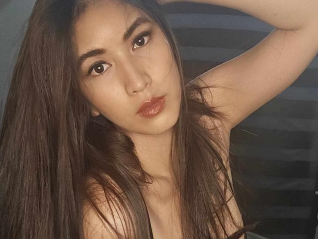 BunnyNikkiTS with long brown hair, dark eyes, and glossy red lips, arm raised, showing bare shoulders, wearing a black top, soft light on face.