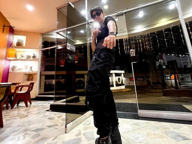 Andrew Carmona, em pé em frente a uma vitrine de vidro, vestindo roupas pretas, óculos de sol e um relógio de pulso, apontando para frente, com uma expressão séria..