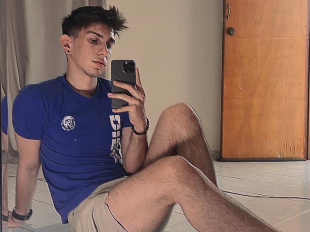 Andrew Carmona, con i capelli castano scuro e arruffati, una maglietta blu e pantaloncini beige, seduto sul pavimento piastrellato, con le gambe incrociate, sta scattando un selfie allo specchio con un telefono nero..