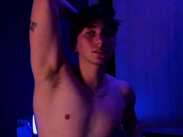 Andrew Carmona senza camicia, con capelli scuri, braccio alzato, che mostra i peli delle ascelle, un piccolo capezzolo, un tatuaggio sulla parte superiore del braccio, illuminazione blu, una leggera espressione facciale, una collana..