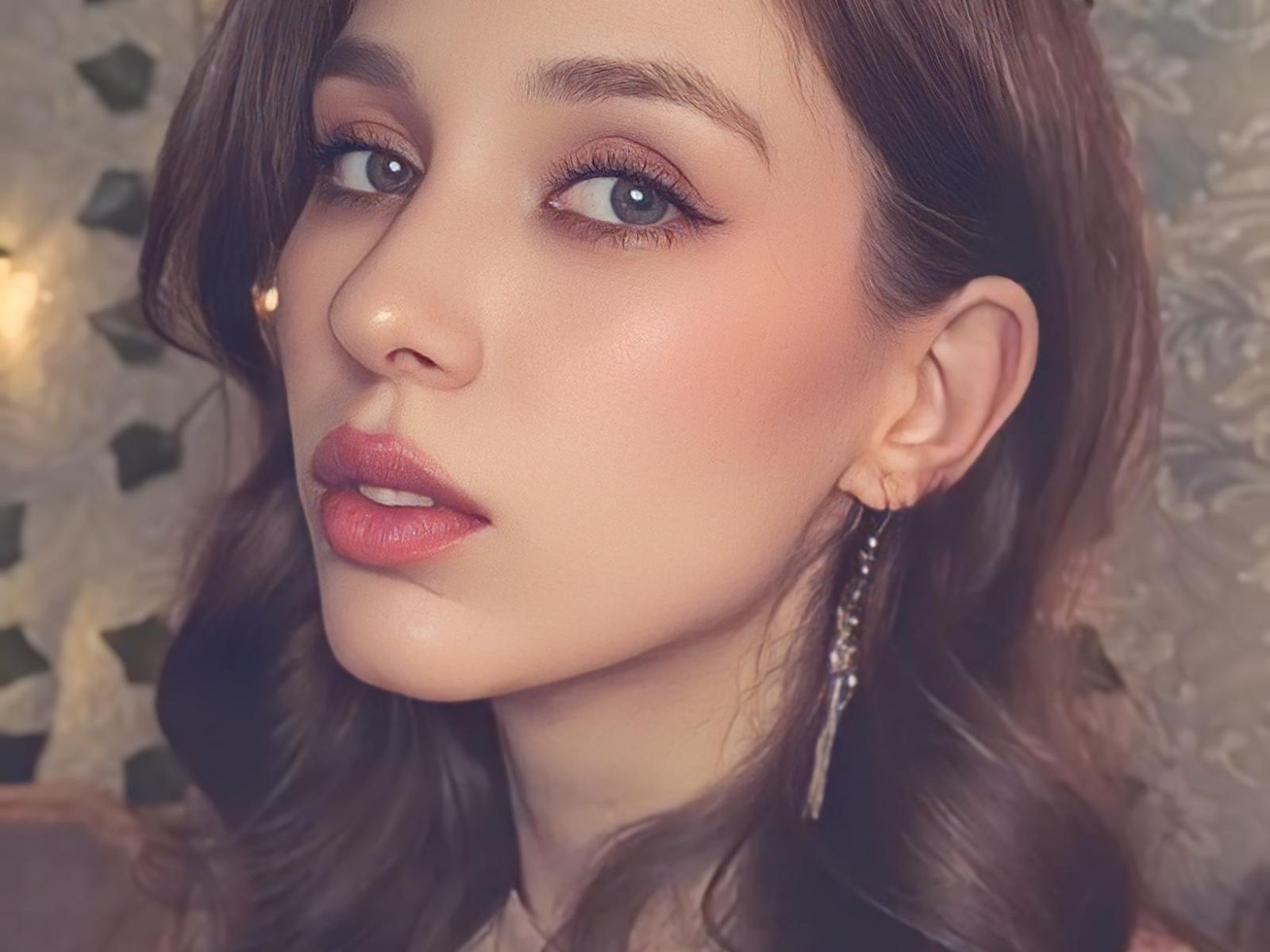 ShawtyVi has pretty brown eyes, pink lipstick, and wavy brown hair with a small nose stud and long, dangling earrings. Her skin is smooth and slightly flushed.